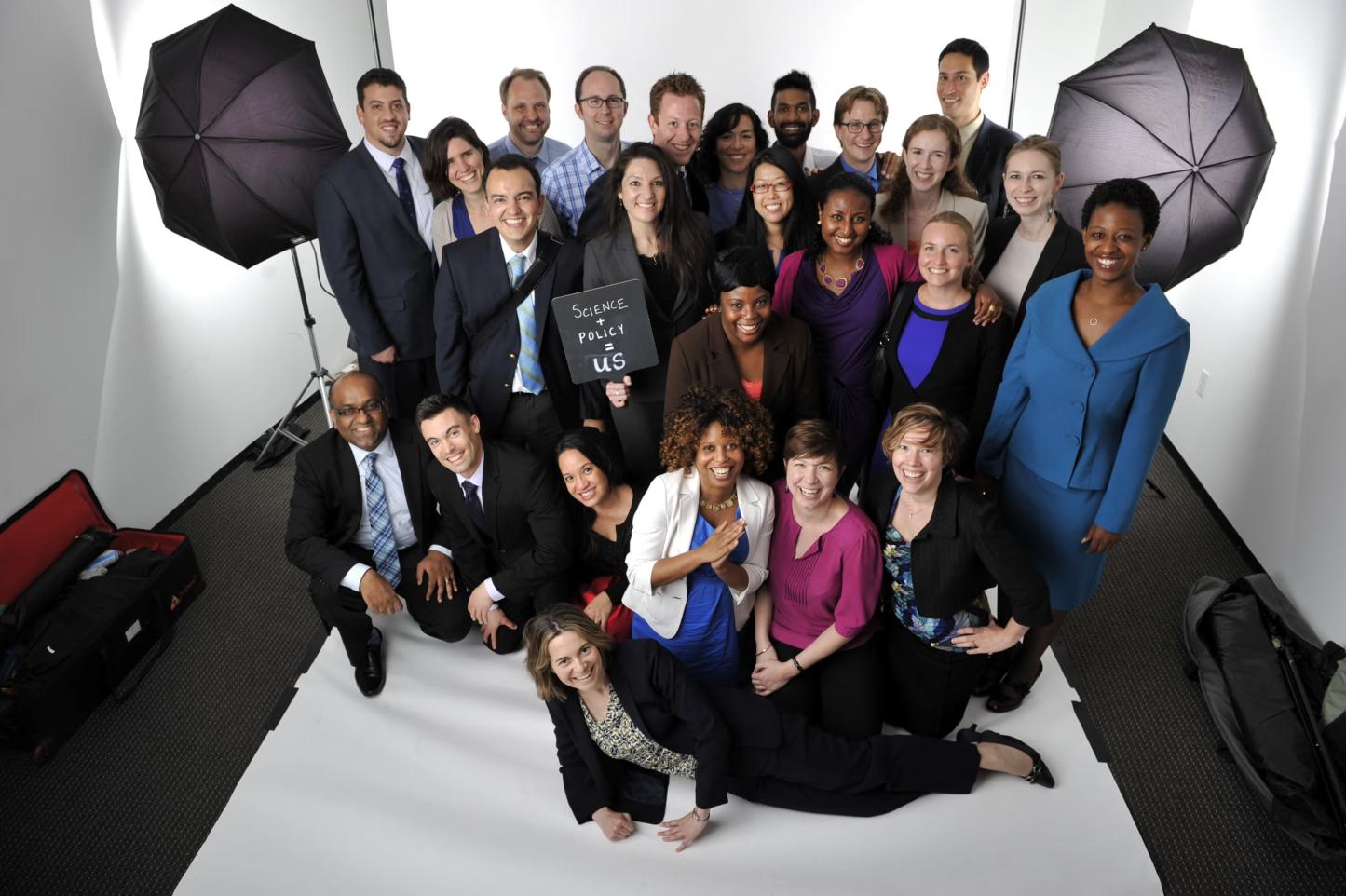 Fellows sitting and smiling and holding a sign that reads science + policy = us.