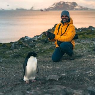 Carrero-Martínez kneeling on the ground at the Henryk Arctowski Polish Antarctic Station, behind a penguin in the foreground.